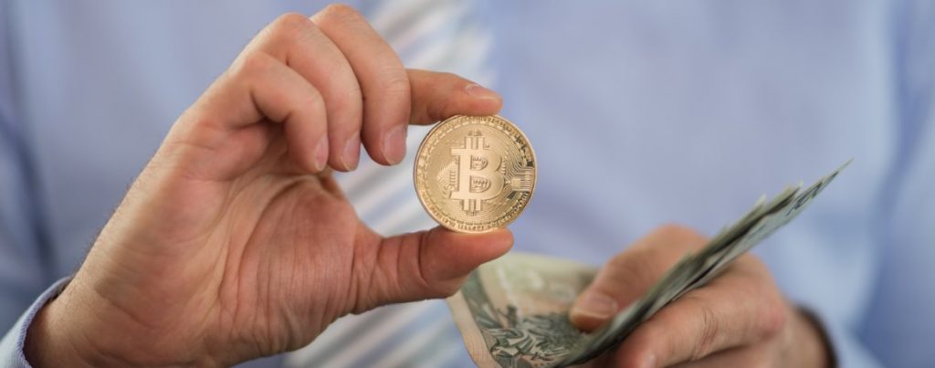 Mann hält eine Bitcoin-Münze neben Geldscheinen – Symbolbild für Kryptowährungen und ihre umsatzsteuerliche Behandlung.