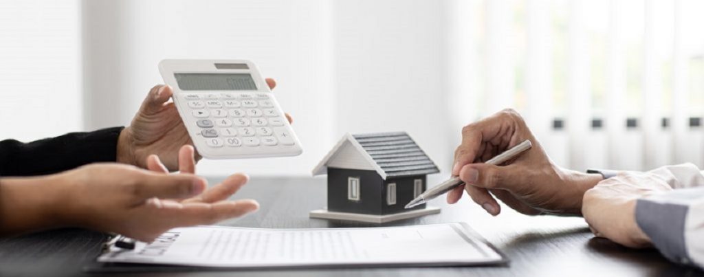 Zwei Personen besprechen steuerliche Aspekte einer vermieteten Immobilie, mit Miniaturhaus, Taschenrechner und Vertragsunterlagen auf dem Tisch.